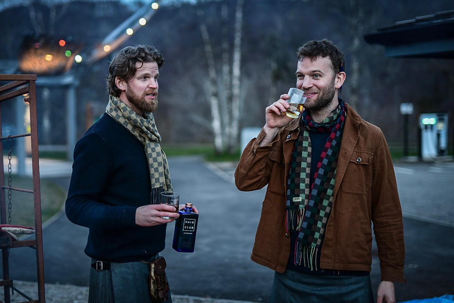 Andy and Calum in kilts with a dram outdoors at dusk