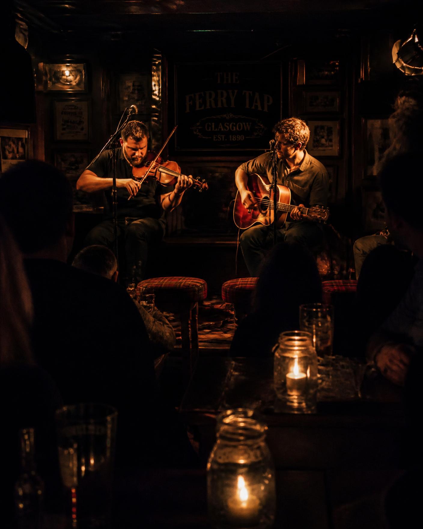 Fiddle and guitar players performing in a dark candlelit bar with tartan stools